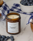 An amber glass jar candle labeled 'Blueberry Muffin' by Sparkle Candle, styled on a white marble surface with blueberries, muffins, and a blue-and-white gingham cloth.
