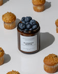 A candle jar labeled 'Blueberry Muffin' by Sparkle Candle, filled with fresh blueberries, styled on a white surface with scattered blueberry muffins around it.