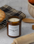 A Sparkle Candle jar candle labeled 'Caramel Apple Pie' sits on a light stone surface with baking props, including a pie, a plaid cloth, wooden utensils, and apples in the background.