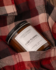 A Sparkle Candle in a clear glass jar with amber-brown wax and a black lid, nestled in a red and black plaid flannel shirt pocket. The label reads 'COZY FLANNEL'.