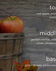 A rustic autumn-themed graphic with a wooden basket holding red apples against a weathered wood background. White text labels color layers: 'top' as red apple, peach, and orange; 'middle' as golden nectar, warm clove, and cinnamon; 'base' as green leaves and tonka bean.