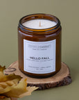 An amber glass jar candle with a white label reading 'SPARKLE CANDLE' and 'HELLO FALL' sits on a rustic wood slice with dried leaves against a muted olive background.