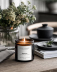 Lit candle in an amber jar with a label on a wooden surface, surrounded by decor items.