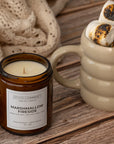 An amber glass jar candle labeled Sparkle Candle Marshmallow Fireside sits on a rustic wooden table beside a beige ceramic mug filled with toasted marshmallows.