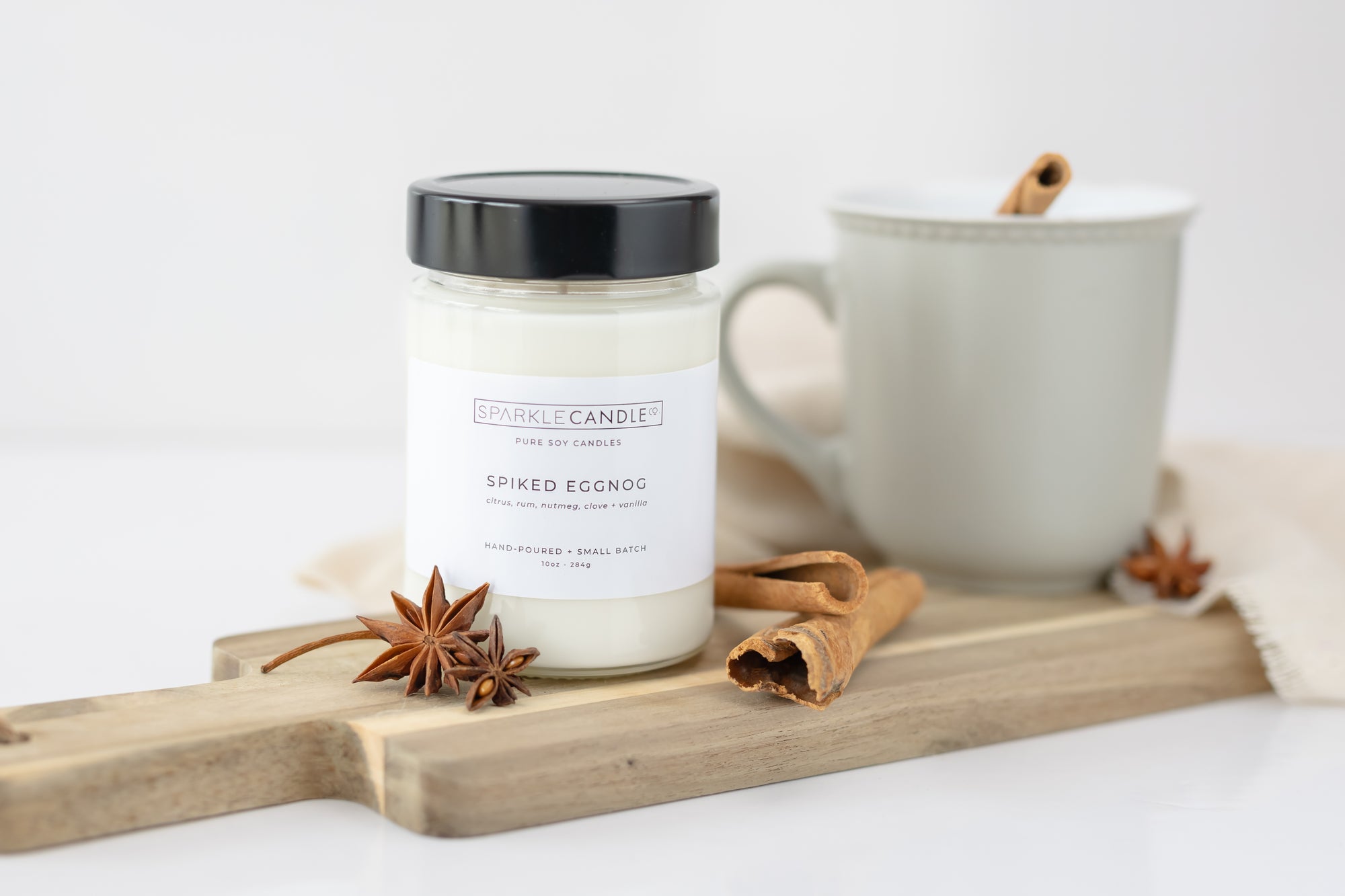 A jar candle with a label reading 'Spiked Eggnog', set on a wooden board alongside a mug of eggnog, spices, and decorative items.
