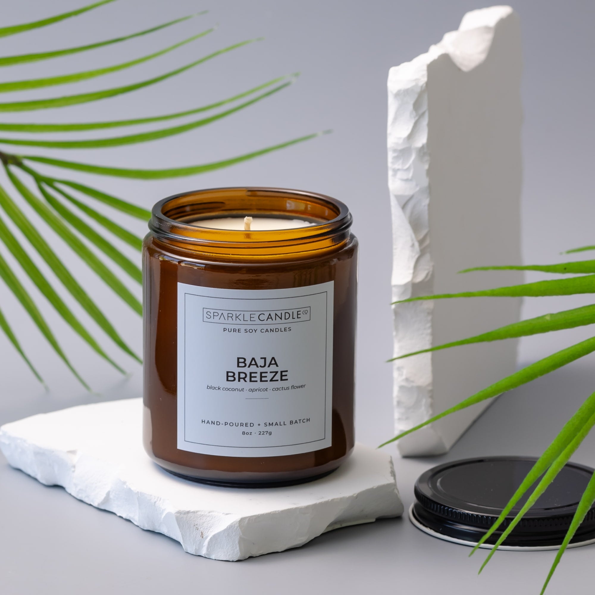Sparkle Candle Baja Breeze candle in an amber jar on a white surface with green leaves in the background