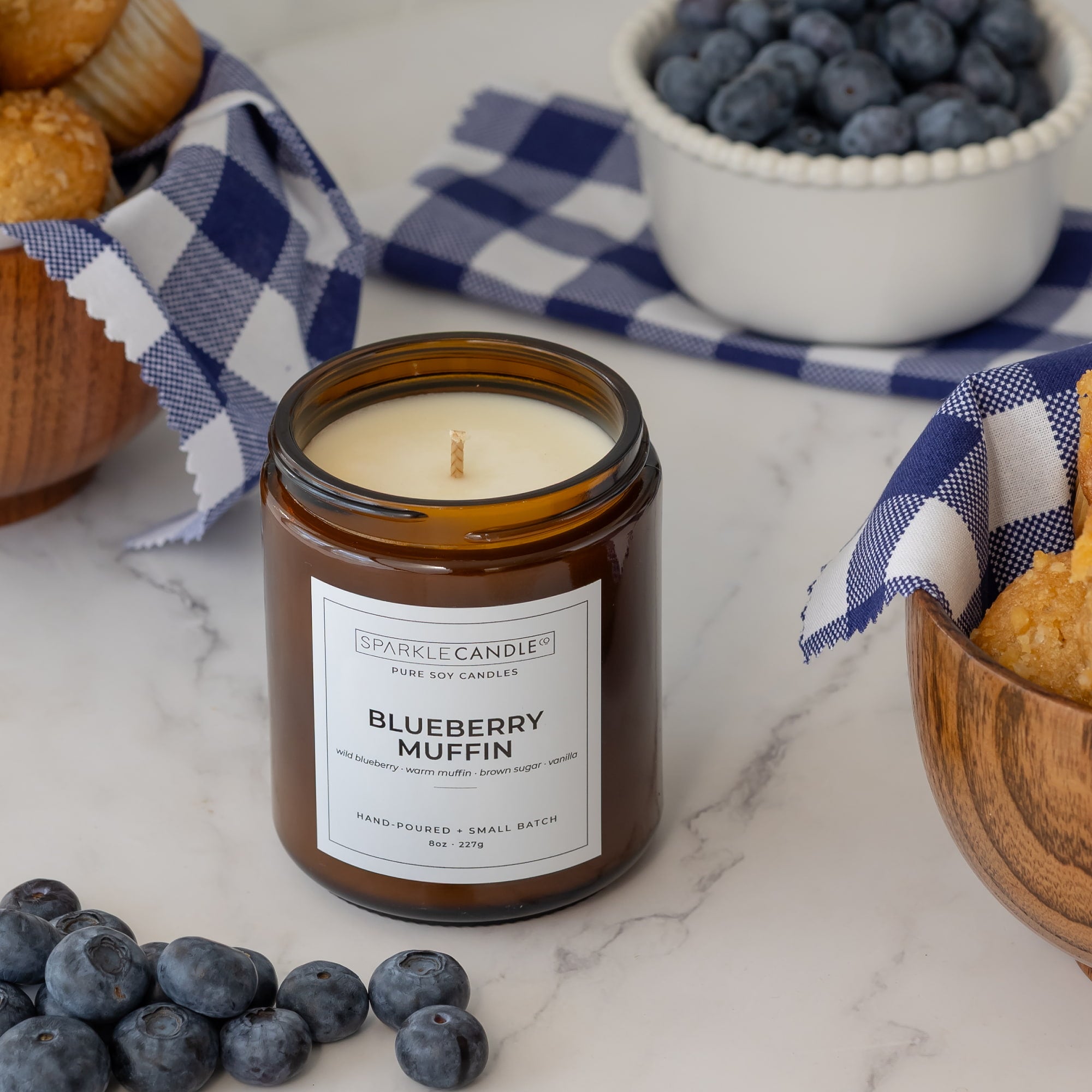 An amber glass jar candle labeled 'Blueberry Muffin' by Sparkle Candle, styled on a white marble surface with blueberries, muffins, and a blue-and-white gingham cloth.