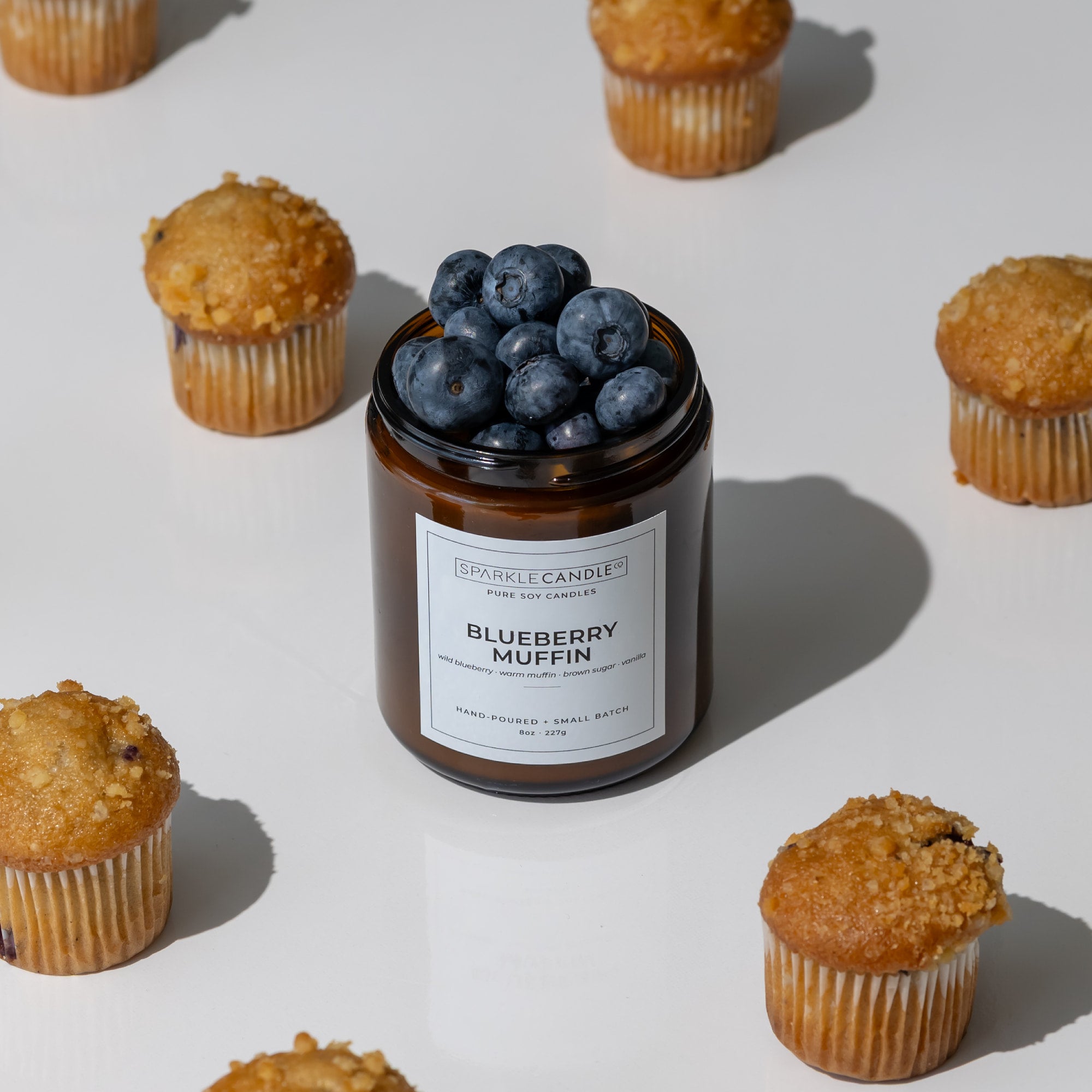 A candle jar labeled 'Blueberry Muffin' by Sparkle Candle, filled with fresh blueberries, styled on a white surface with scattered blueberry muffins around it.