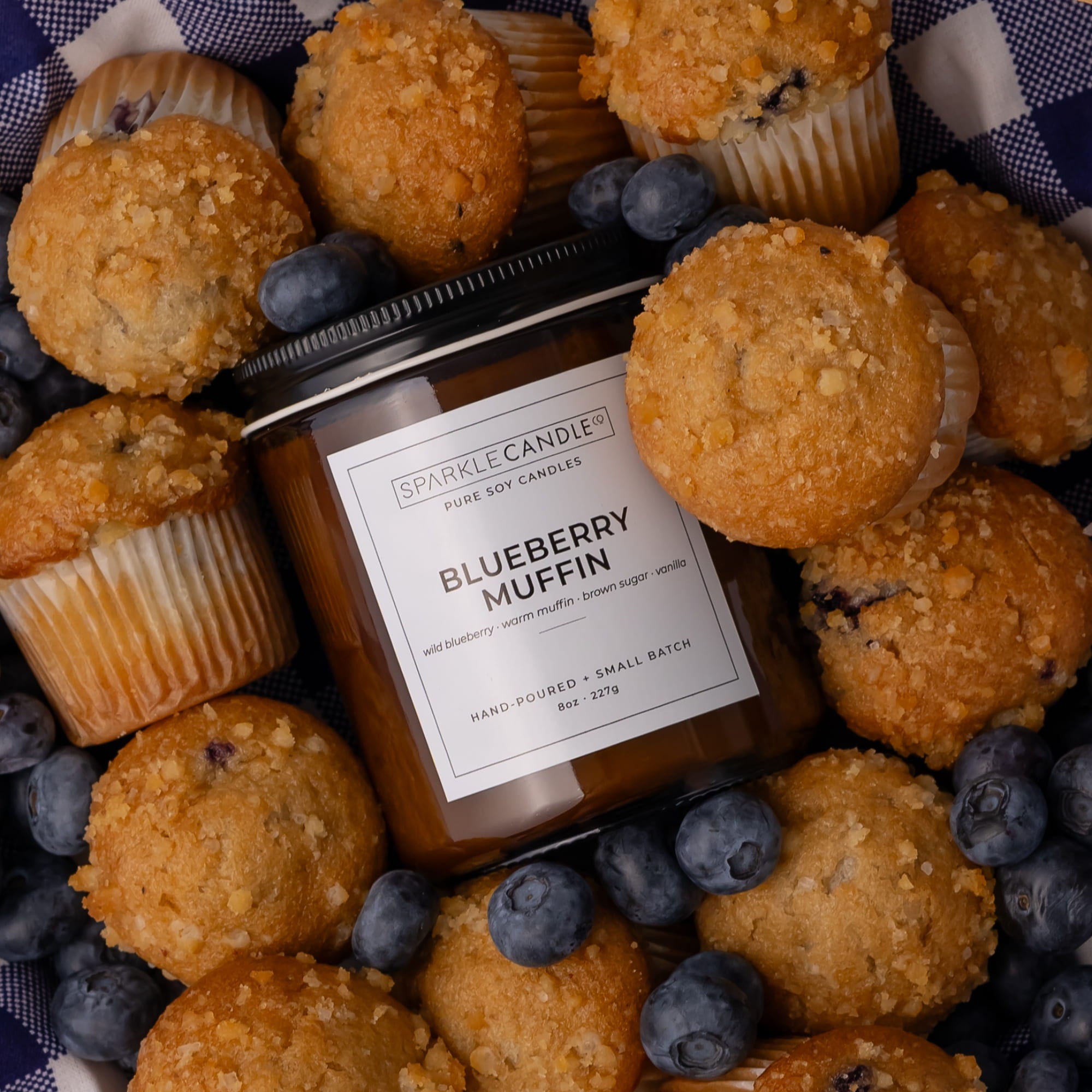 A jar candle labeled Sparkle Candle Blueberry Muffin scent, surrounded by blueberry muffins and fresh blueberries on a blue-and-white check fabric background.