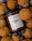 A jar candle labeled Sparkle Candle Blueberry Muffin scent, surrounded by blueberry muffins and fresh blueberries on a blue-and-white check fabric background.