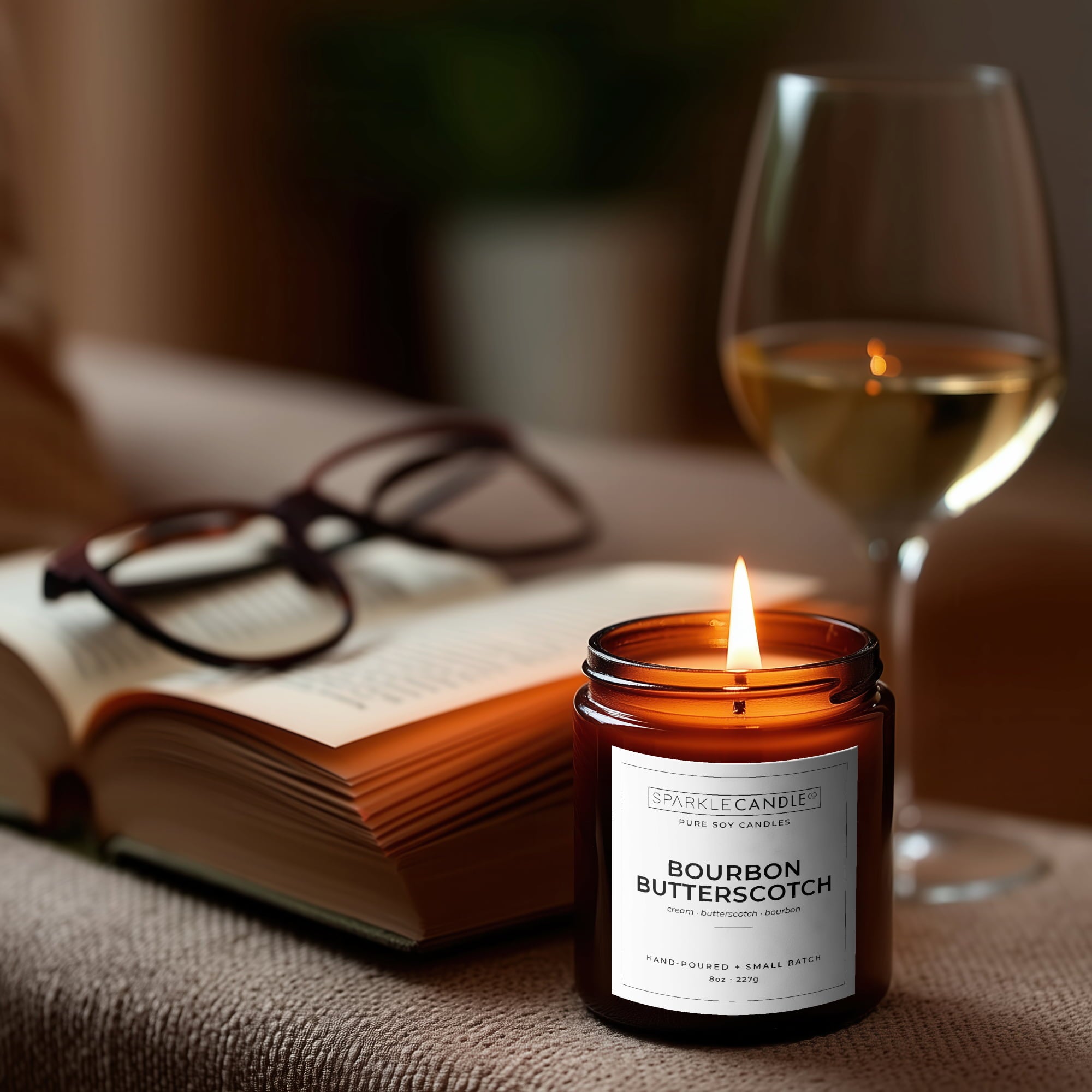 Candle labeled 'Bourbon Butterscotch' next to an open book and a glass of wine on a table.