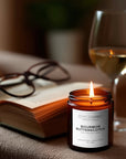 Candle labeled 'Bourbon Butterscotch' next to an open book and a glass of wine on a table.