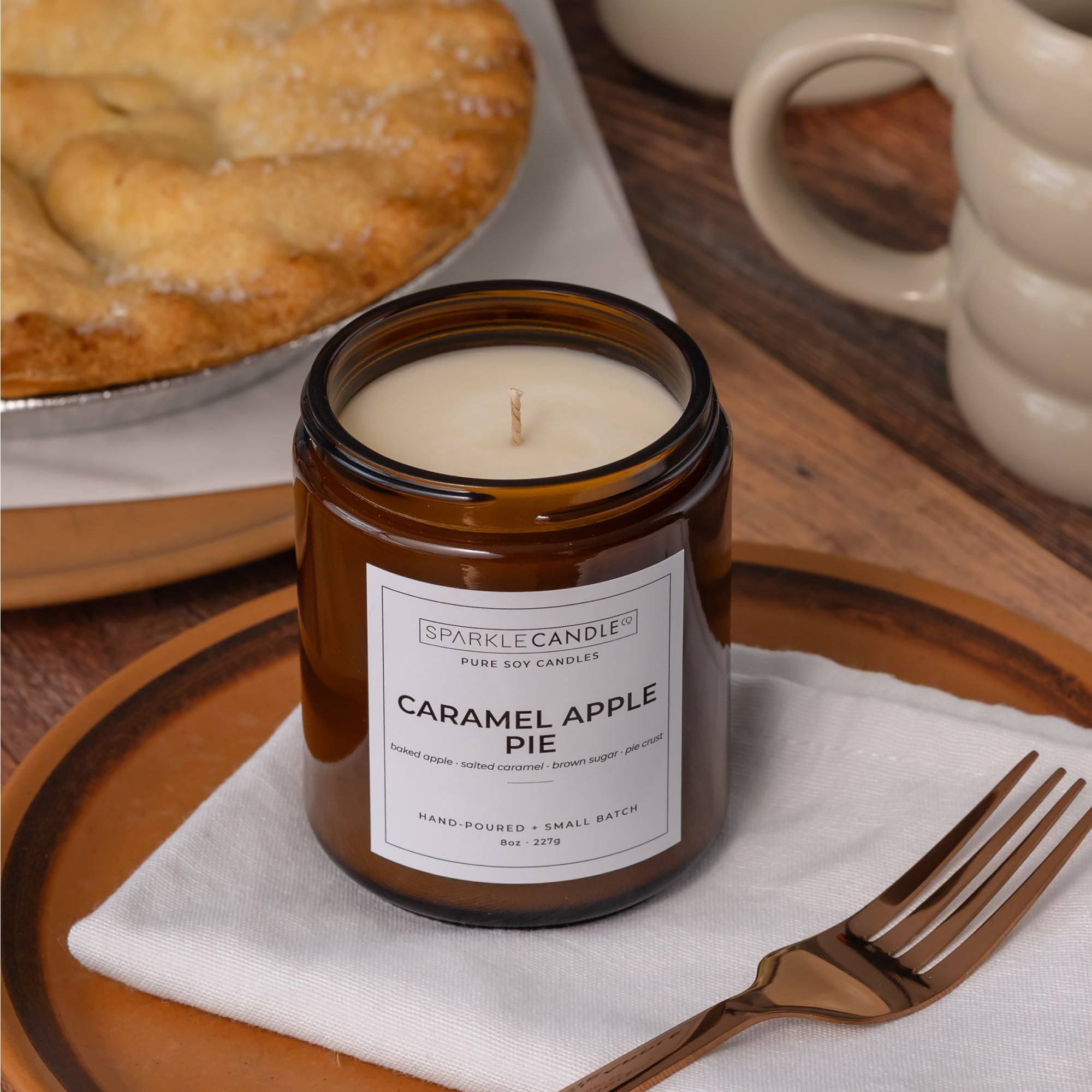 Sparkle Candle Caramel Apple Pie scented soy candle in an amber glass jar, styled on a wooden table with a white napkin, gold fork, and a baked pie in the background.