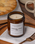 Sparkle Candle Caramel Apple Pie scented soy candle in an amber glass jar, styled on a wooden table with a white napkin, gold fork, and a baked pie in the background.