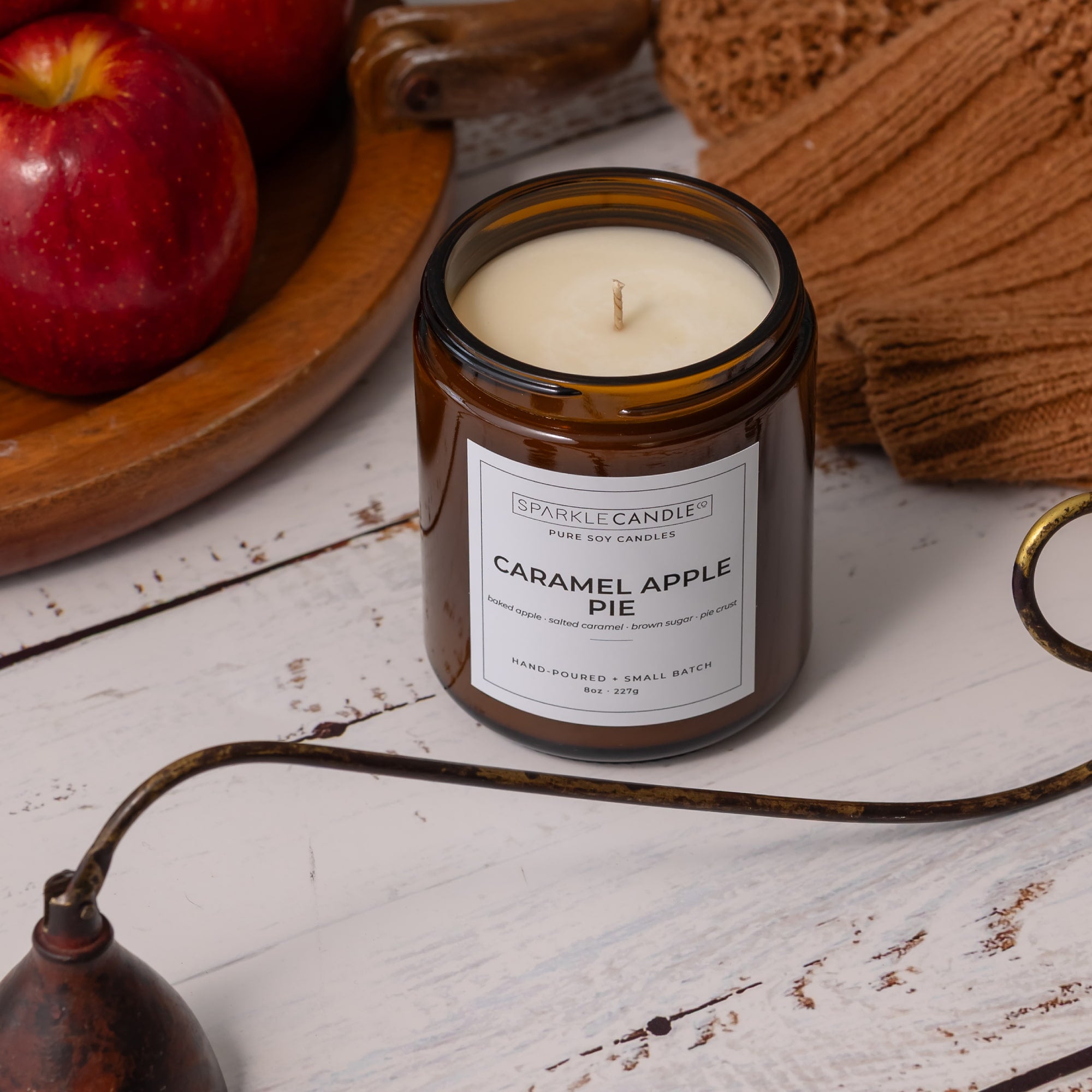 A Sparkle Candle in a small amber glass jar labeled 'Caramel Apple Pie,' styled on a white wood surface with red apples, a brown knit textile, and a brass candle wick holder nearby.