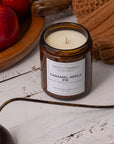 A Sparkle Candle in a small amber glass jar labeled 'Caramel Apple Pie,' styled on a white wood surface with red apples, a brown knit textile, and a brass candle wick holder nearby.