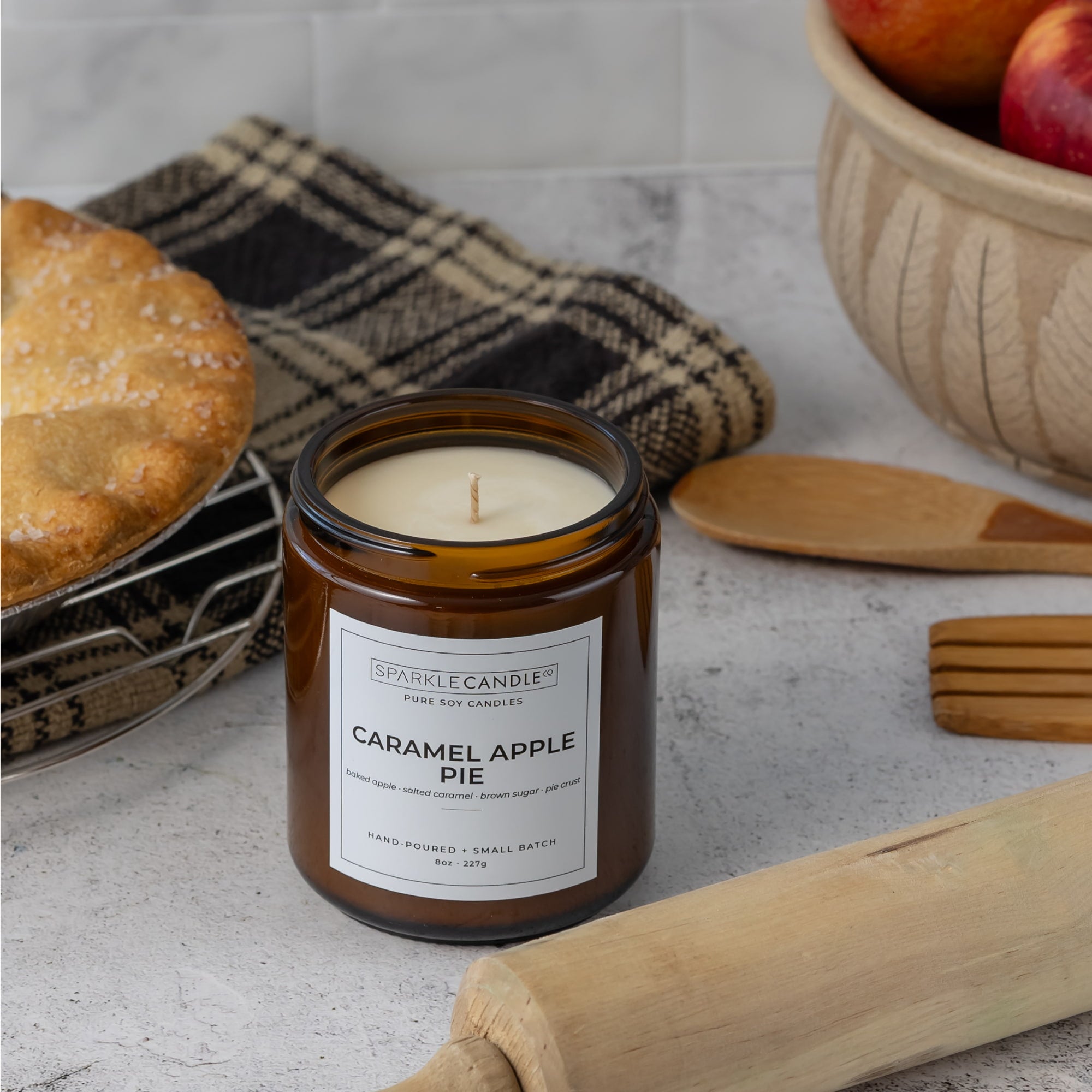 A Sparkle Candle jar candle labeled 'Caramel Apple Pie' sits on a light stone surface with baking props, including a pie, a plaid cloth, wooden utensils, and apples in the background.