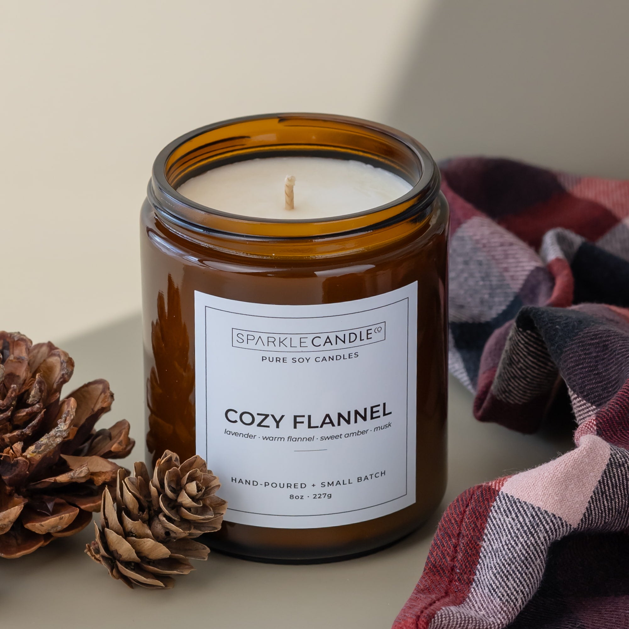 An amber glass jar candle with a white label reading 'SPARKLE CANDLE' and 'COZY FLANNEL' is styled on a beige surface with pinecones and a plaid blanket in the background.