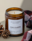 An amber glass jar candle with a white label reading 'SPARKLE CANDLE' and 'COZY FLANNEL' is styled on a beige surface with pinecones and a plaid blanket in the background.