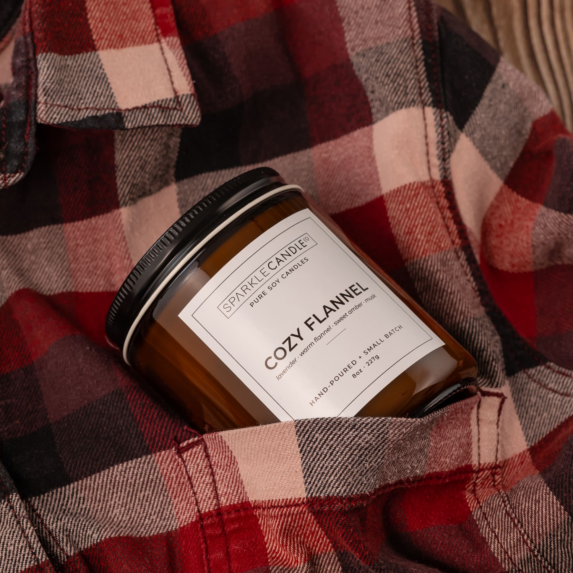 A Sparkle Candle in a clear glass jar with amber-brown wax and a black lid, nestled in a red and black plaid flannel shirt pocket. The label reads 'COZY FLANNEL'.