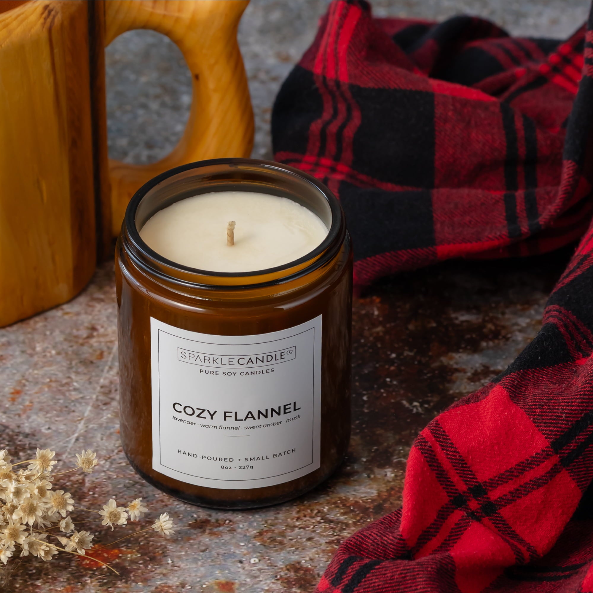 An amber glass jar candle labeled Sparkle Candle Cozy Flannel, styled on a rustic stone surface with a red and black plaid blanket, a wooden mug, and dried flowers.