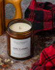 An amber glass jar candle labeled Sparkle Candle Cozy Flannel, styled on a rustic stone surface with a red and black plaid blanket, a wooden mug, and dried flowers.