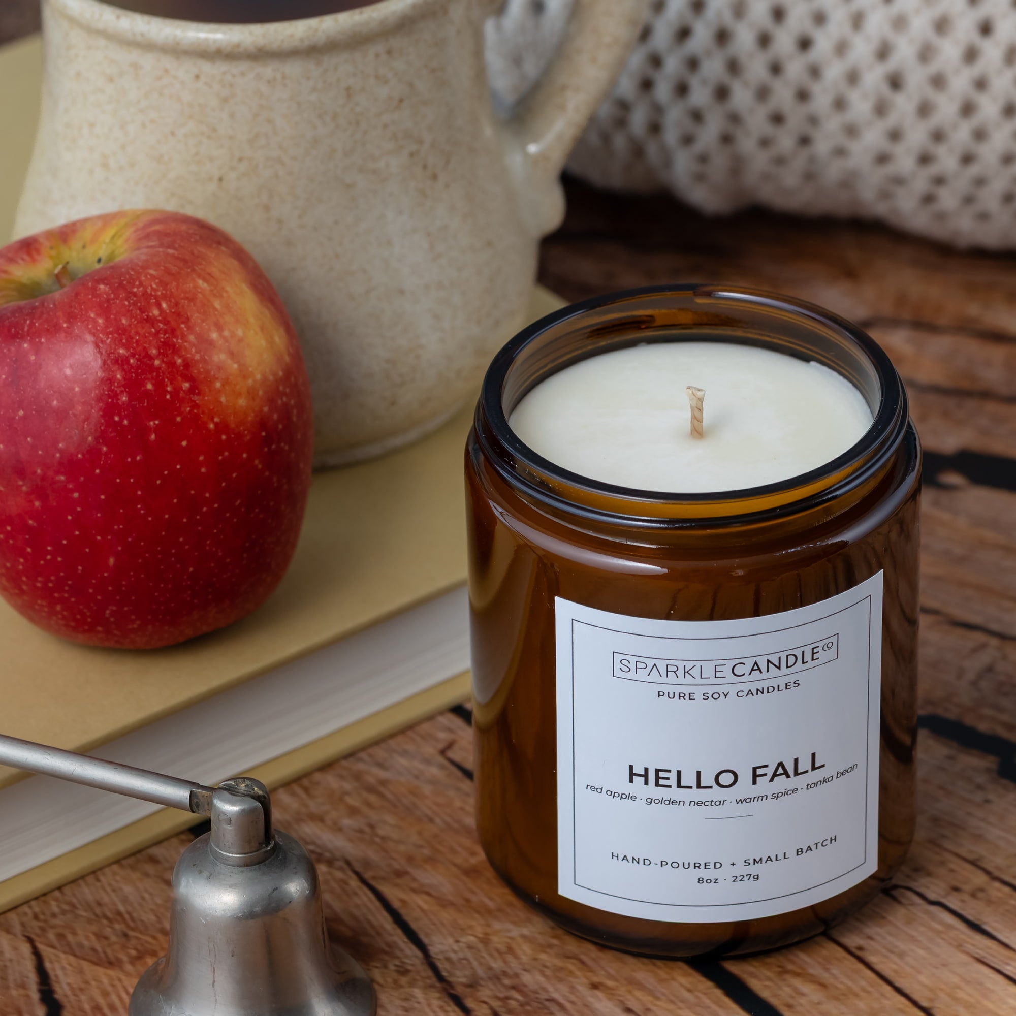 An amber glass jar candle labeled 'HELLO FALL' by Sparkle Candle sits on a rustic wooden surface, styled with a red apple, a ceramic mug, and a metal bell.