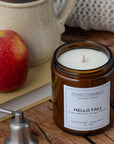 An amber glass jar candle labeled 'HELLO FALL' by Sparkle Candle sits on a rustic wooden surface, styled with a red apple, a ceramic mug, and a metal bell.