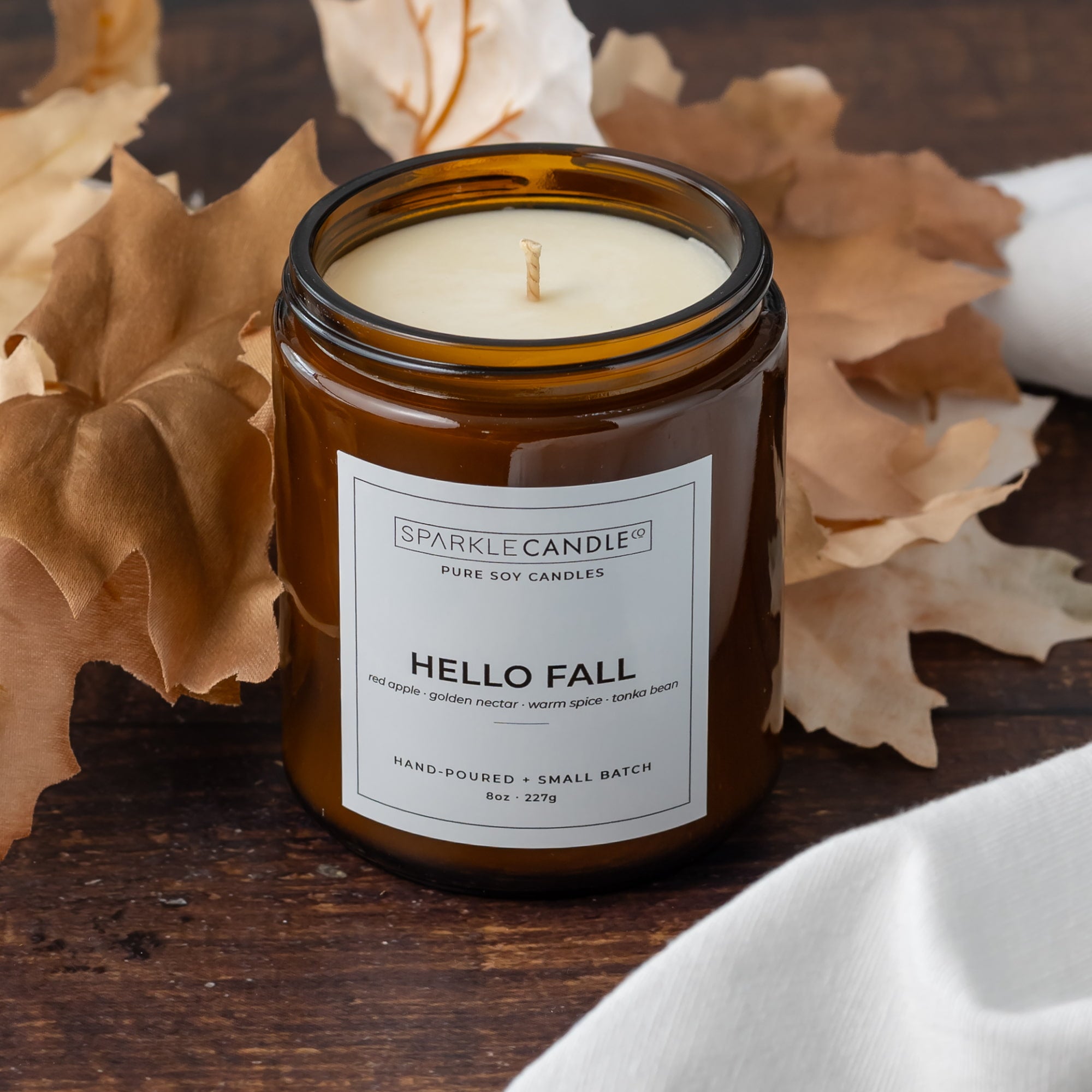 An amber glass jar candle labeled 'Sparkle Candle Co. Hello Fall' sits on a rustic wooden surface surrounded by dried autumn leaves and a white cloth.