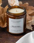 An amber glass jar candle labeled 'Sparkle Candle Co. Hello Fall' sits on a rustic wooden surface surrounded by dried autumn leaves and a white cloth.