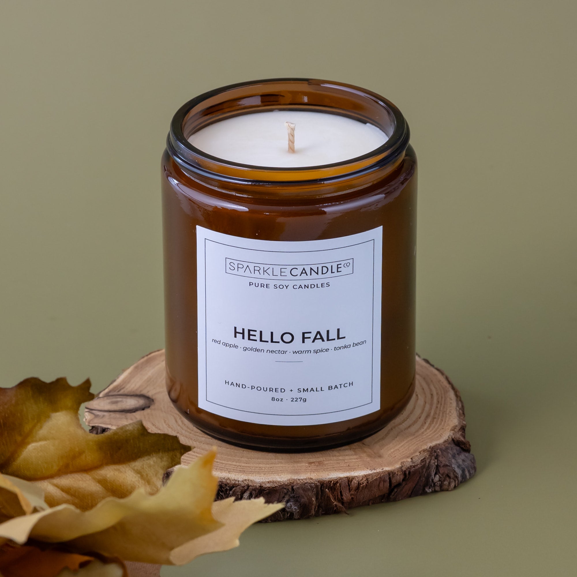 An amber glass jar candle with a white label reading 'SPARKLE CANDLE' and 'HELLO FALL' sits on a rustic wood slice with dried leaves against a muted olive background.