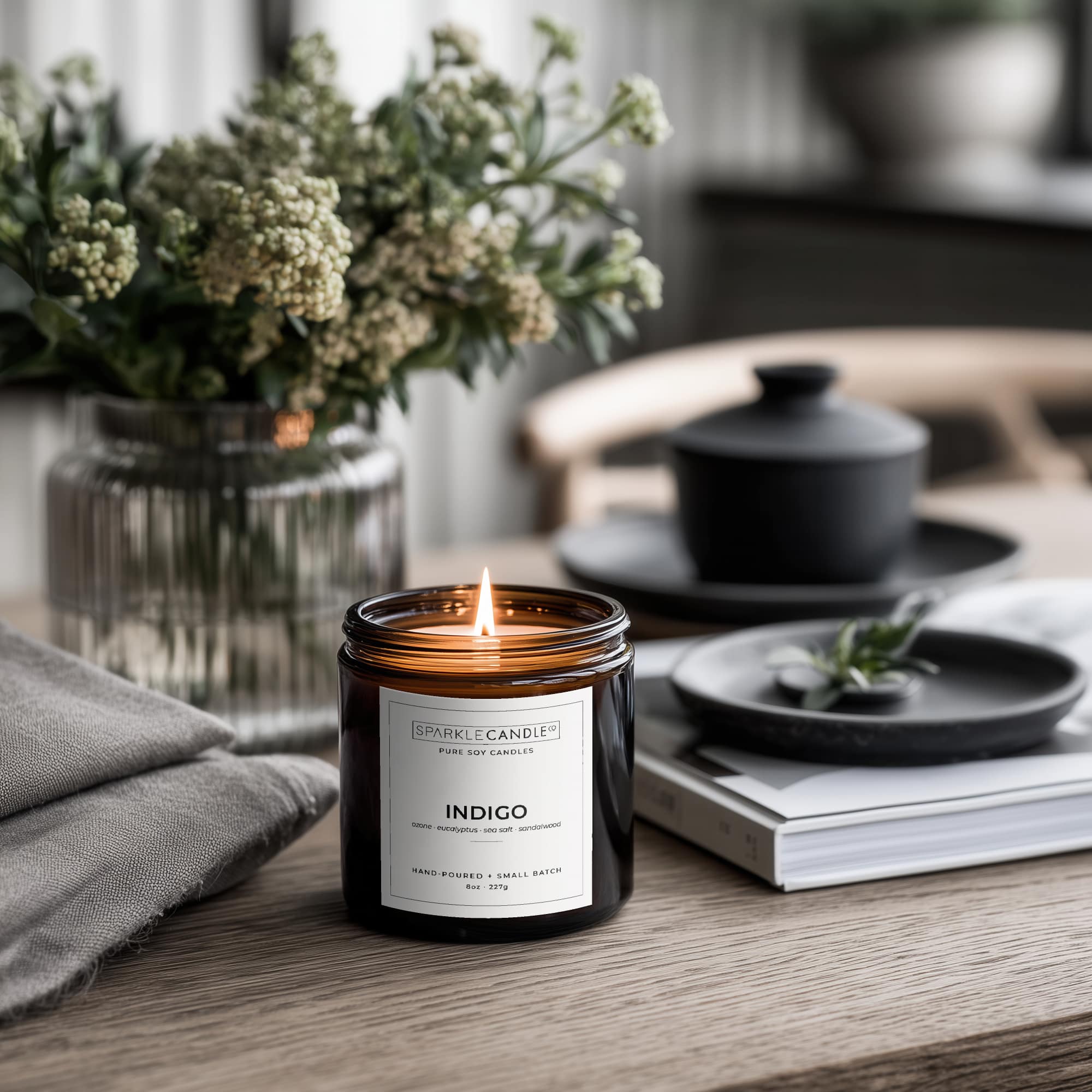 Lit candle in an amber jar with a label on a wooden surface, surrounded by decor items.