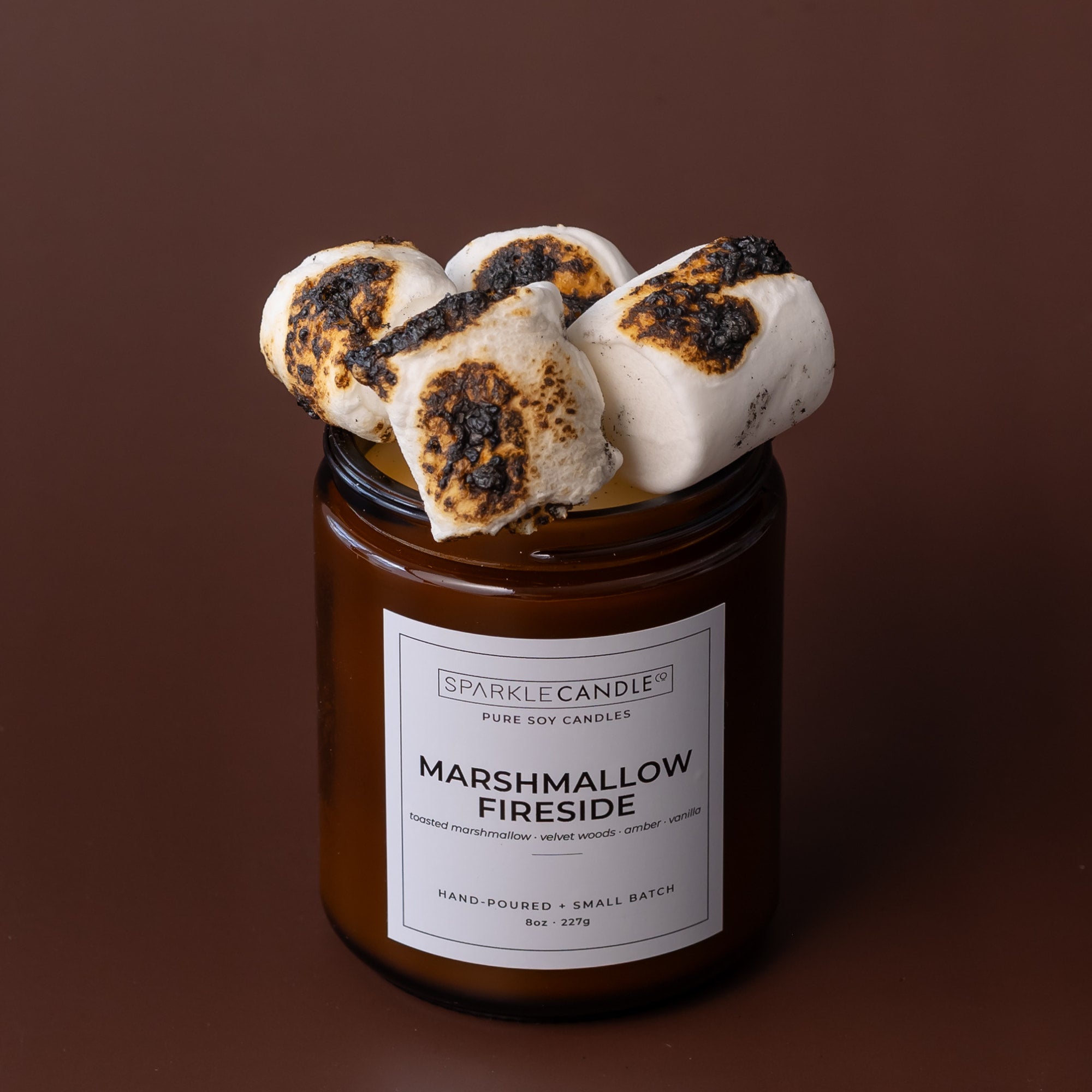 An amber glass jar candle with a white label reading Sparkle Candle Marshmallow Fireside, topped with three toasted marshmallows with charred spots, set against a solid dark brown background.