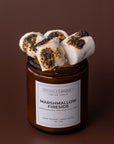 An amber glass jar candle with a white label reading Sparkle Candle Marshmallow Fireside, topped with three toasted marshmallows with charred spots, set against a solid dark brown background.