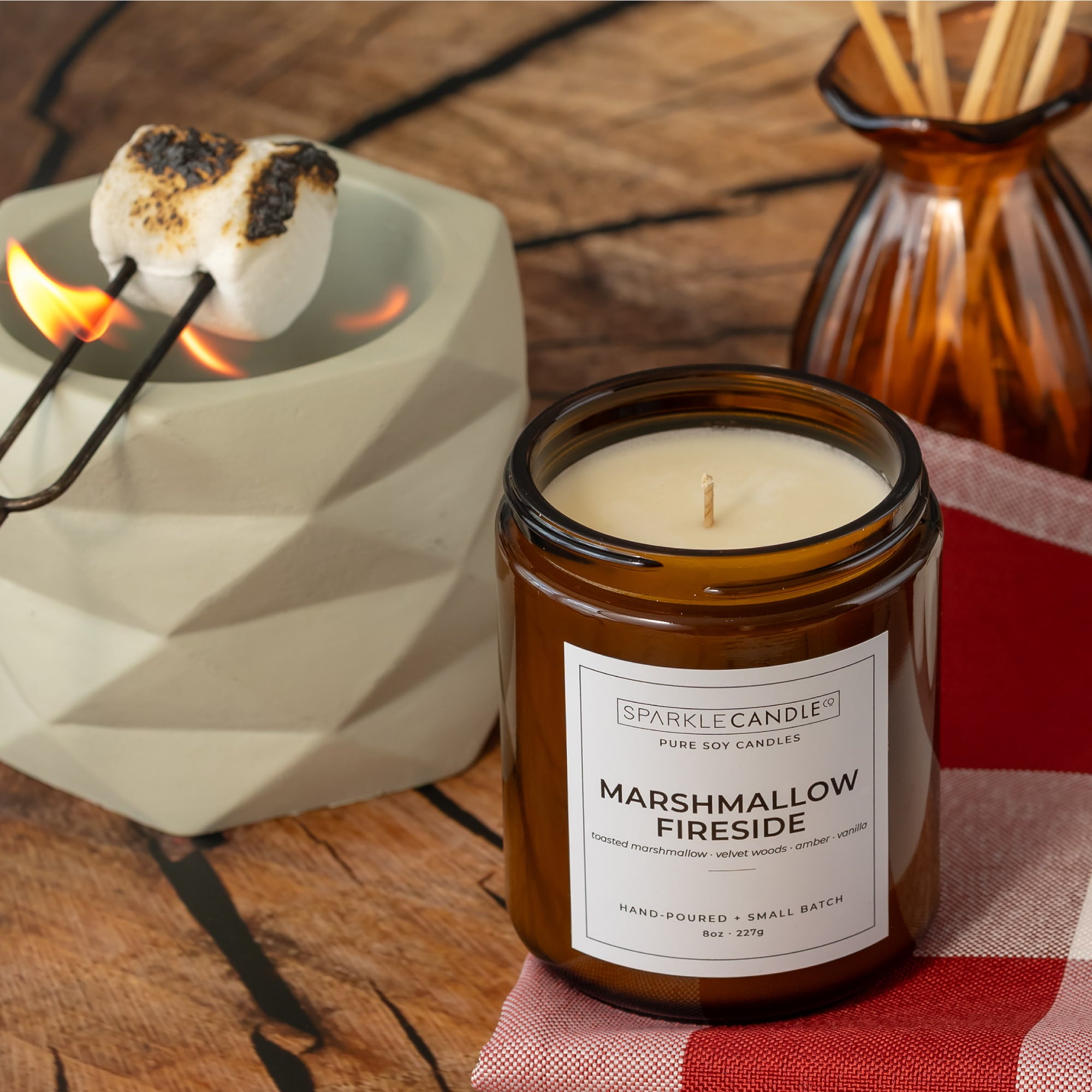 An amber glass jar candle labeled Sparkle Candle Marshmallow Fireside sits on a rustic wooden surface beside a white geometric dish holding a toasted marshmallow over a flame, with a brown reed diffuser in the background.