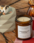 An amber glass jar candle labeled Sparkle Candle Marshmallow Fireside sits on a rustic wooden surface beside a white geometric dish holding a toasted marshmallow over a flame, with a brown reed diffuser in the background.