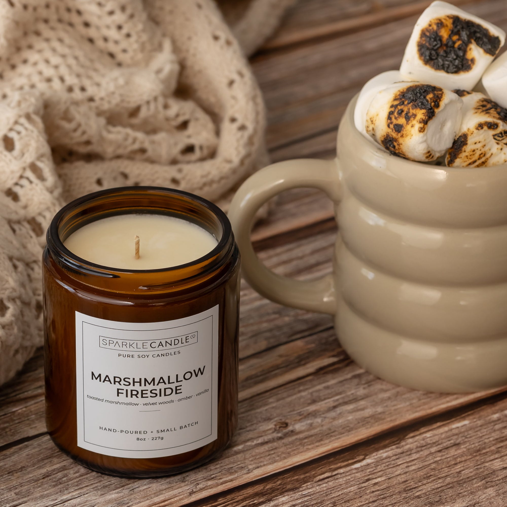 An amber glass jar candle labeled Sparkle Candle Marshmallow Fireside sits on a rustic wooden table beside a beige ceramic mug filled with toasted marshmallows.