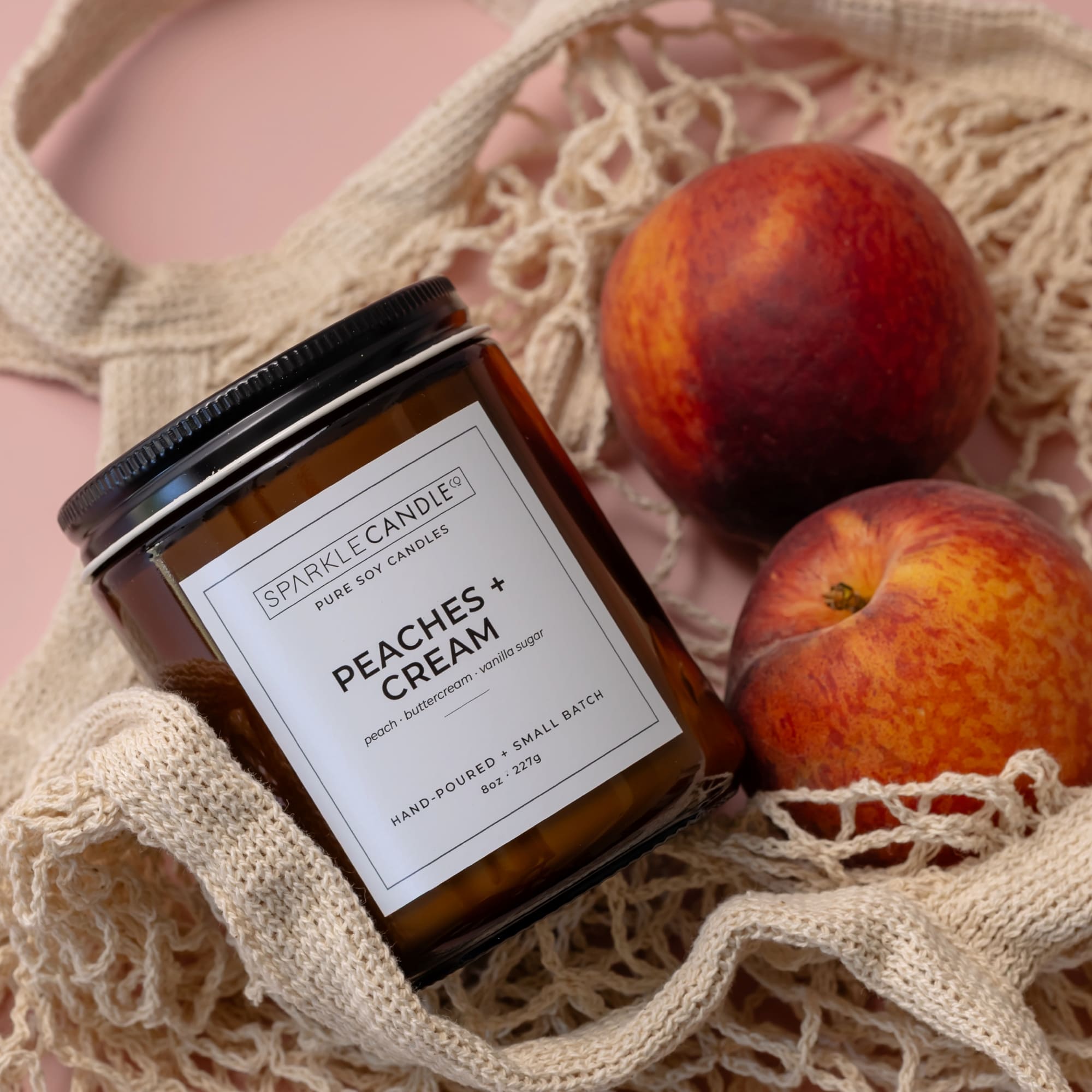 Sparkle Candle 'Peaches + Cream' candle in a mesh bag with peaches on a pink background