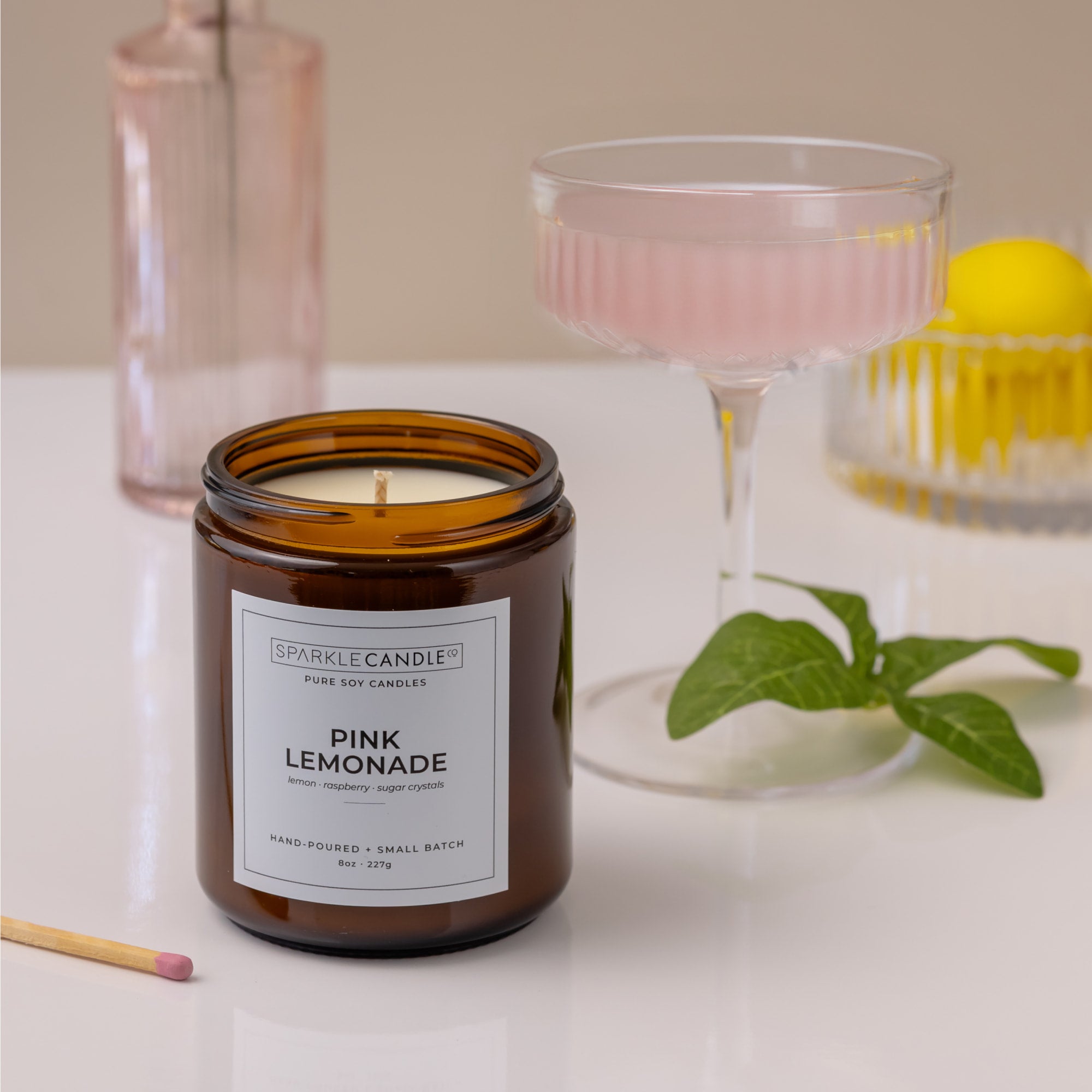 Candle labeled 'Pink Lemonade' with a glass drink container and lemon in the background.