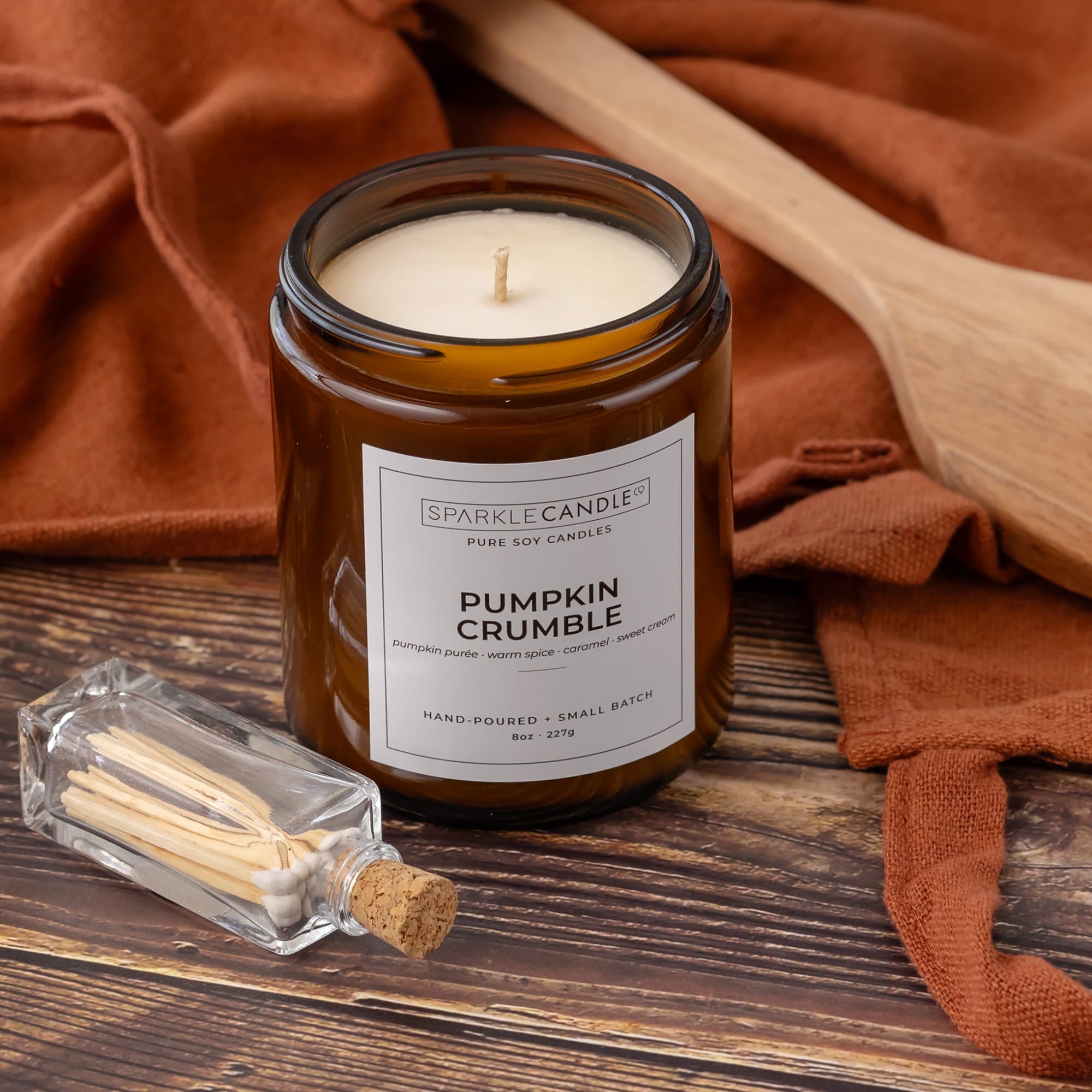 A small amber glass jar candle labeled 'Pumpkin Crumble' by Sparkle Candle, placed on a rustic wooden surface with rust-colored fabric and a small glass match bottle nearby.