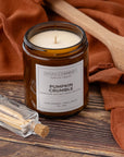 A small amber glass jar candle labeled 'Pumpkin Crumble' by Sparkle Candle, placed on a rustic wooden surface with rust-colored fabric and a small glass match bottle nearby.
