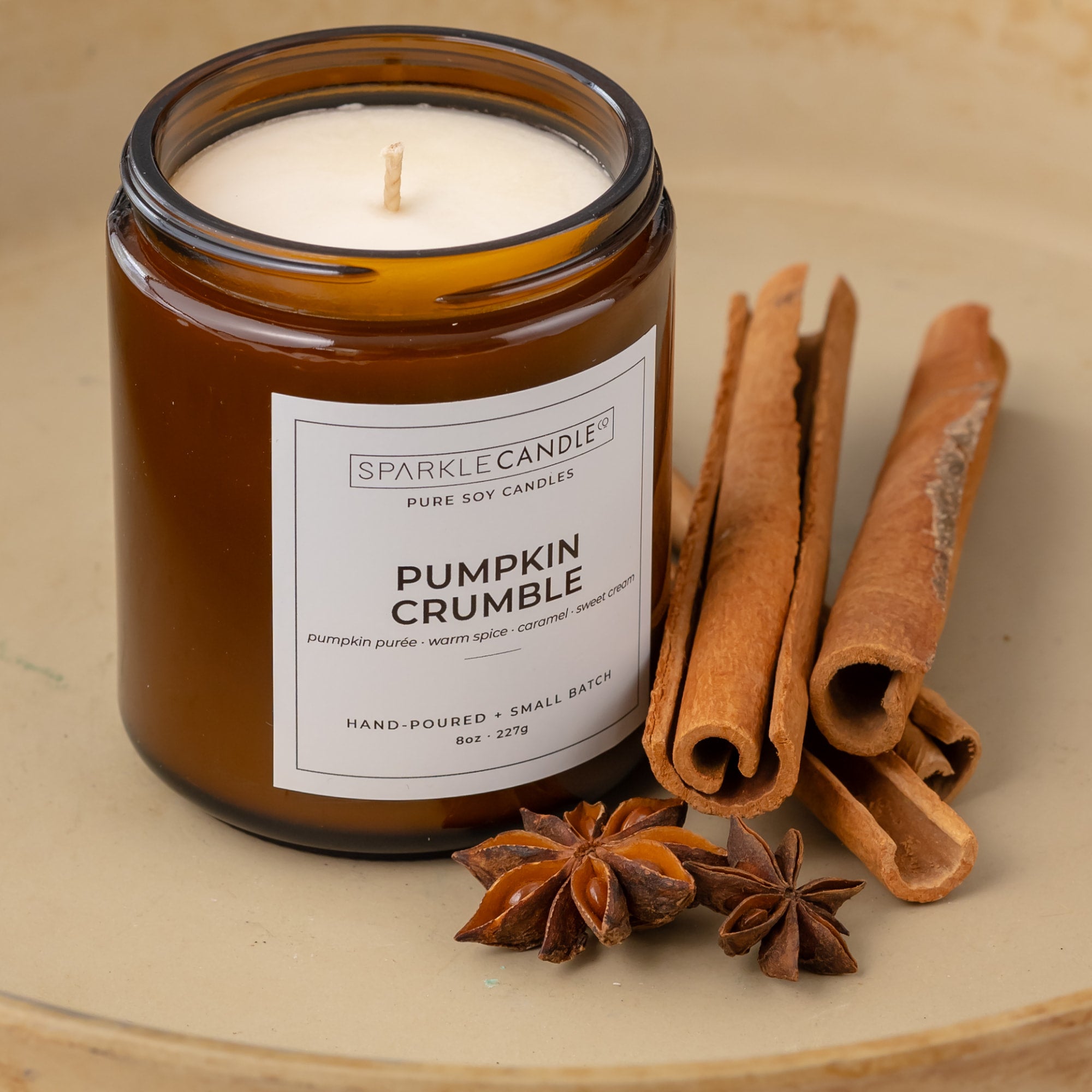 Sparkle Candle 'Pumpkin Crumble' soy candle in an amber glass jar, styled with cinnamon sticks and star anise on a beige ceramic surface.