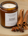 Sparkle Candle 'Pumpkin Crumble' soy candle in an amber glass jar, styled with cinnamon sticks and star anise on a beige ceramic surface.