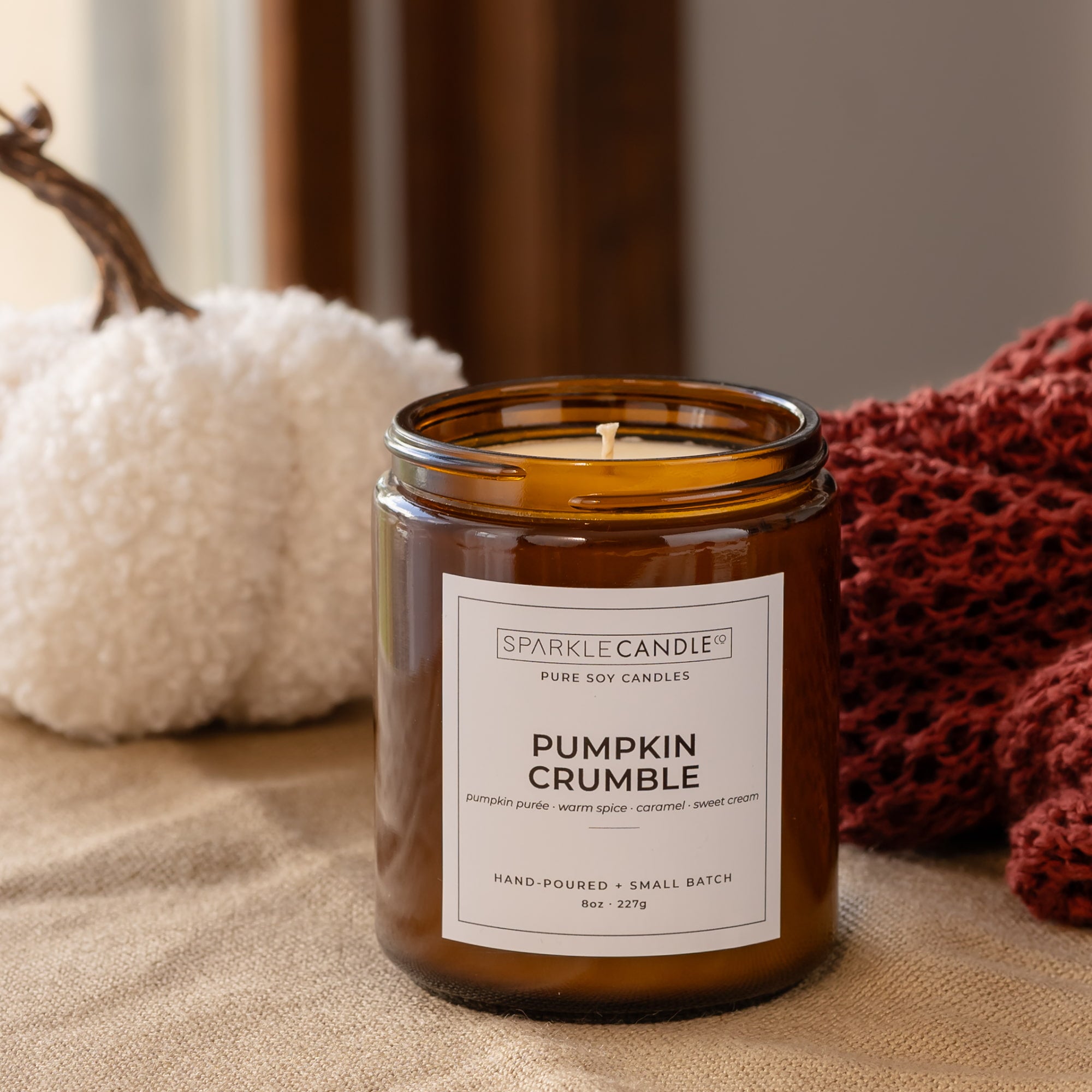 An amber glass jar candle labeled 'Pumpkin Crumble' by Sparkle Candle Co., styled on a linen surface with a white pumpkin and a red knit blanket in the background.