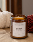 An amber glass jar candle labeled 'Pumpkin Crumble' by Sparkle Candle Co., styled on a linen surface with a white pumpkin and a red knit blanket in the background.