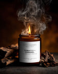 Candle labeled 'Tobacco + Caramel' with smoke and wooden elements on a dark background