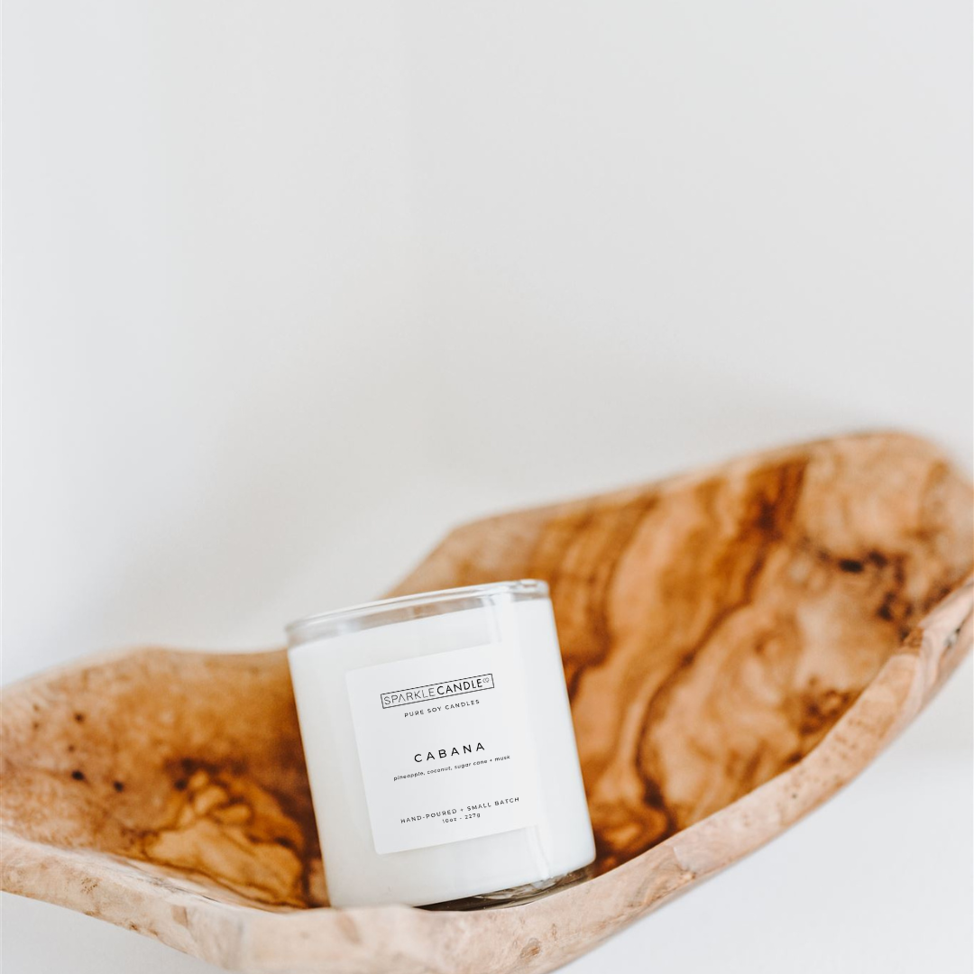 A white candle labeled 'Cabana' with a scent of pineapple, coconut, sugarcane, and musk, placed inside a wooden bowl.